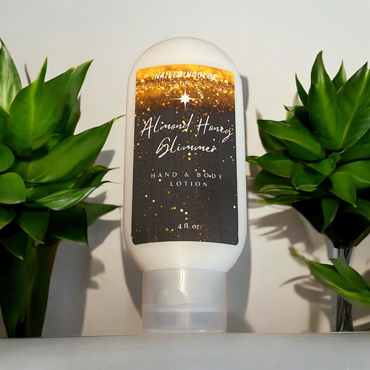 A white squeeze bottle of Hand & Body Lotion labeled 'Almond Honey Glimmer with a gold glittery label, set on a white surface between two green potted plants
