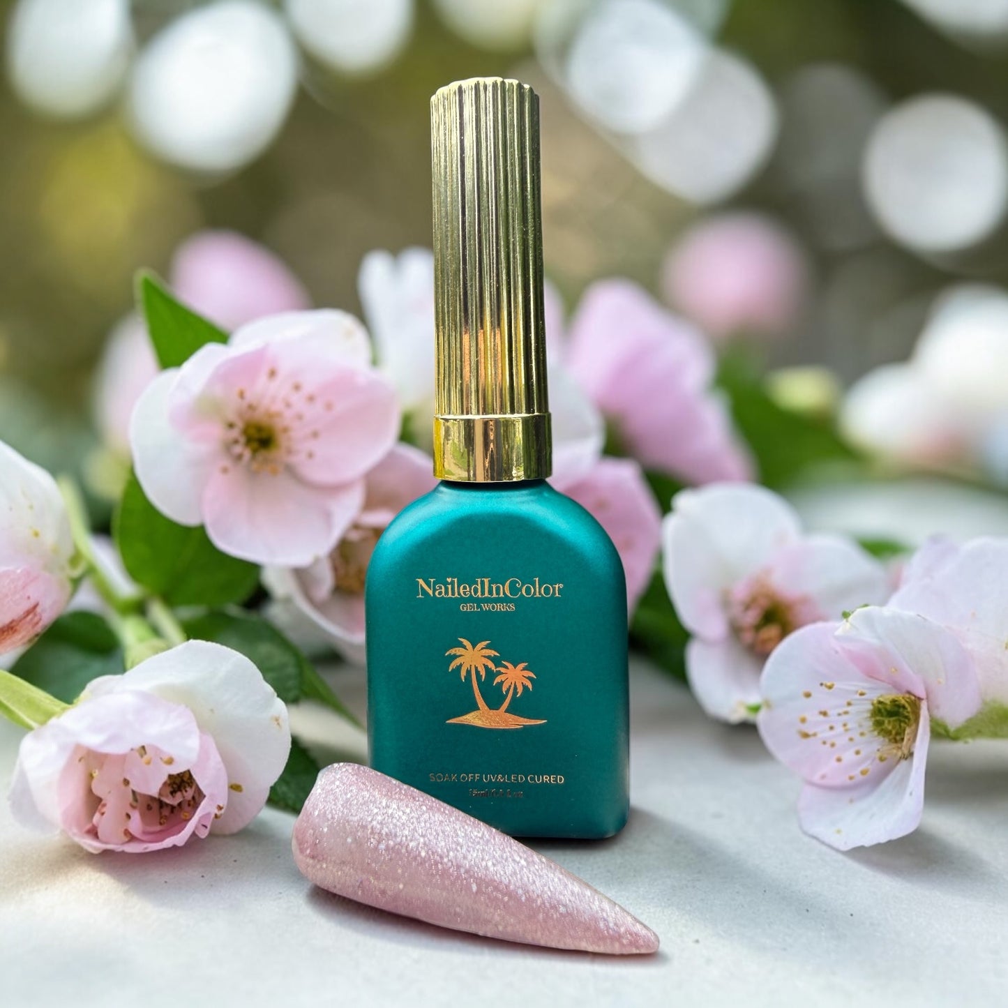 NailedInColor nail polish bottle with a gold cap on a white surface with glitter lavender swatch and flowers in the background