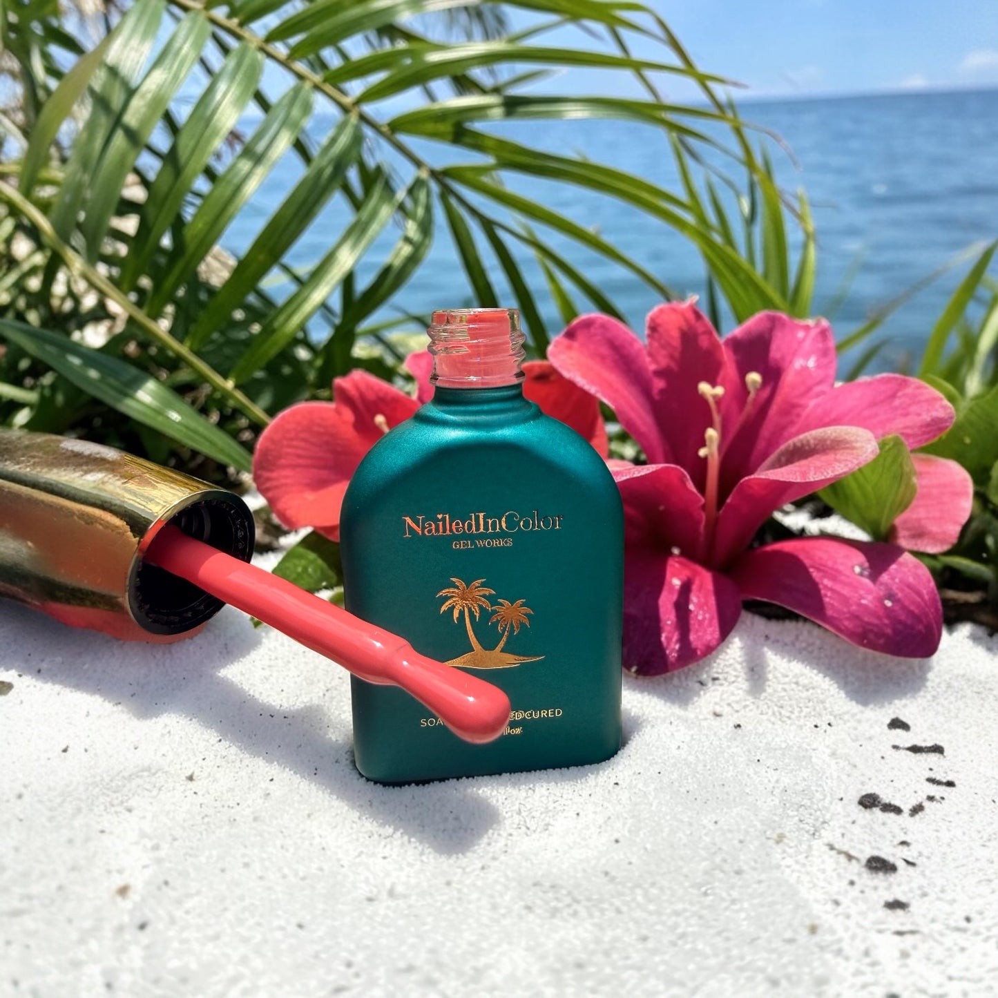 NailedInColor nail polish bottle with brush applicator showing coral gel polish on a sandy surface with flowers and ocean view