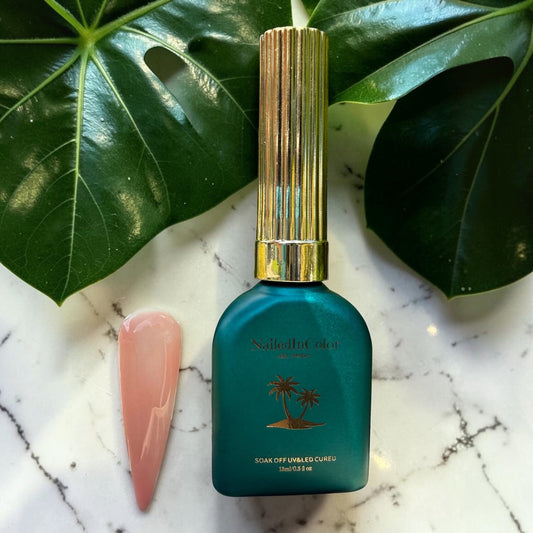 Teal nail polish bottle with gold cap on a marble surface with green leaves in the background and a light pink, beige nail tip in builder gel