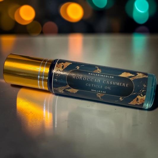 A bottle of NailedinColor Moroccan Cashmere Cuticle Oil with a gold cap and a dark blue label with gold patterns, resting on a reflective metallic surface with colorful bokeh lights in the background.