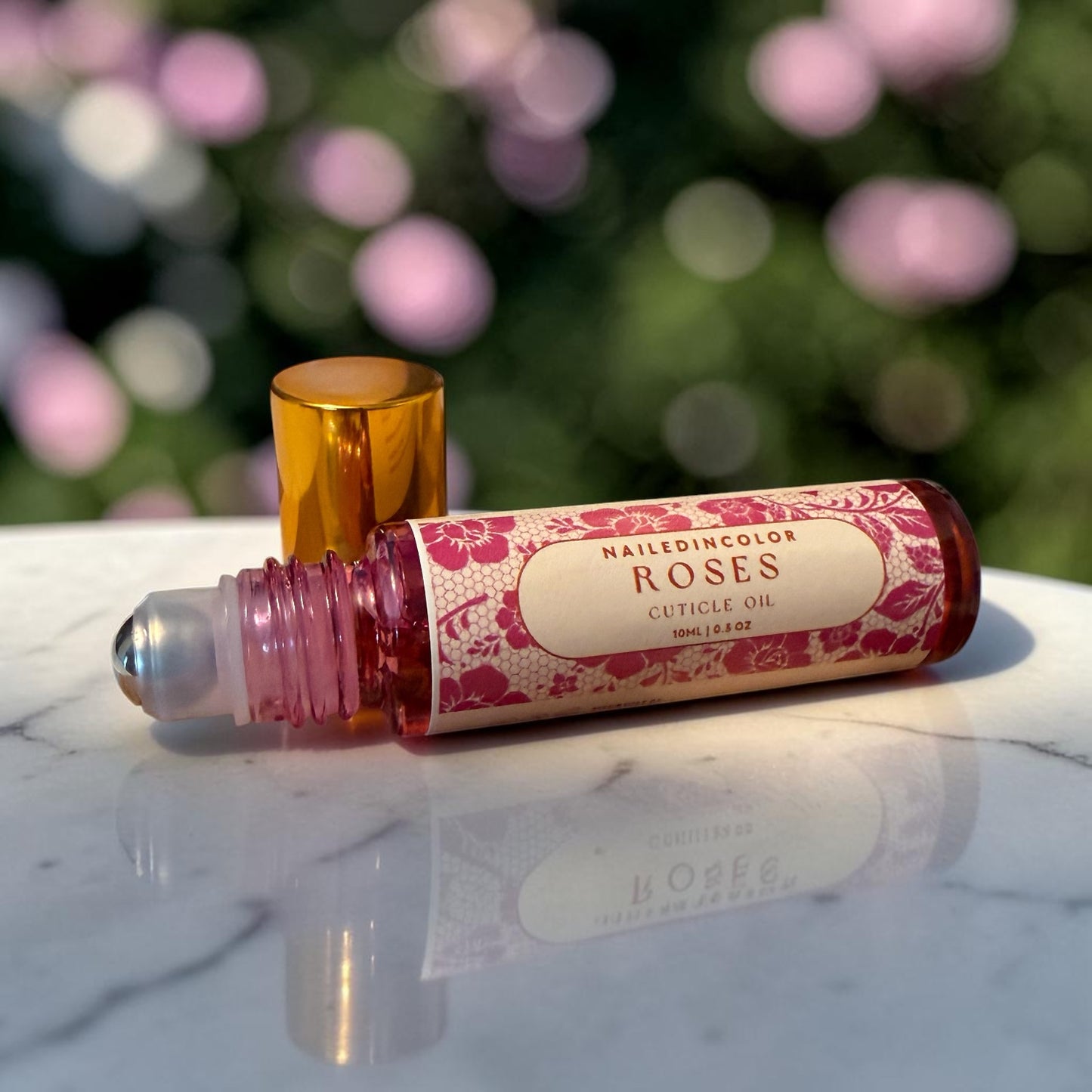 A roll-on bottle of NailedinColor Roses Cuticle Oil with its gold cap removed, resting on a white marble surface with a blurred background of pink flowers.