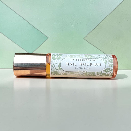 A roll-on bottle of NailedinColor Nail Nourish Cuticle Oil with a metallic rose gold cap and a white floral label, resting on a white surface against a mint green geometric background.