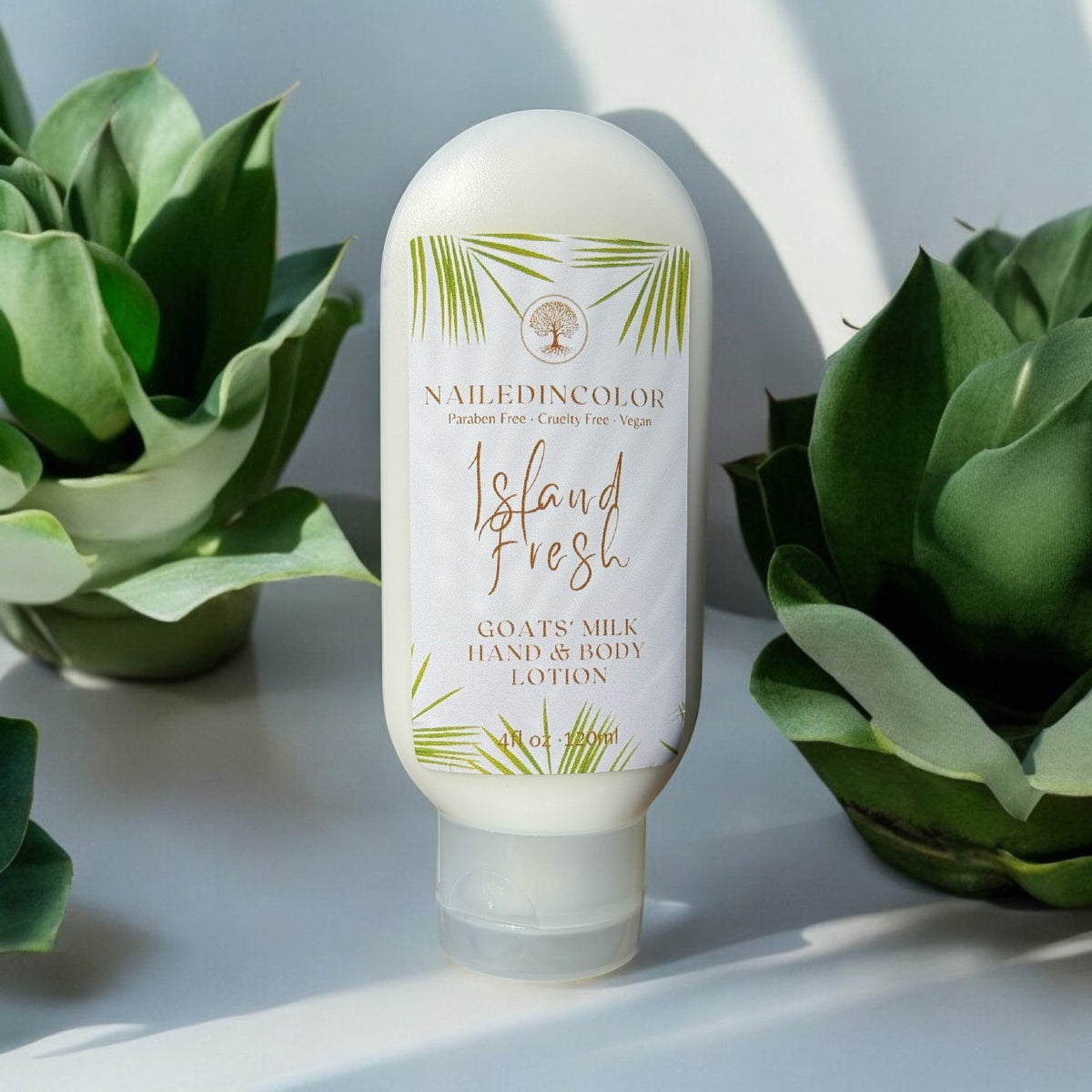 A bottle of NailedinColor 'Island Fresh' goats' milk hand and body lotion stands on a white surface between two green succulent plants, lit by soft natural light with a shadow.