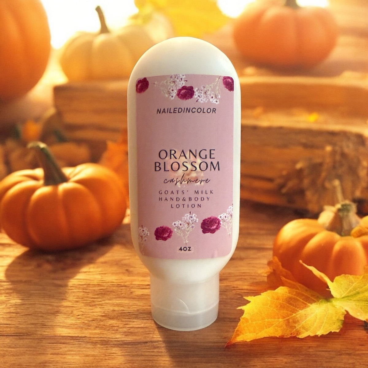 A 4oz bottle of NailedinColor Orange Blossom Cashmere Hand & Body Lotion stands on a wooden surface surrounded by pumpkins and autumn leaves. The bottle is white with a light pink label featuring purple floral illustrations.