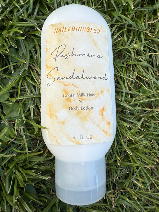 A bottle of NailedinColor Pashmina Sandalwood Body Lotion resting on green grass. The bottle is white with a cream and gold marble-patterned label and a clear flip-top cap.