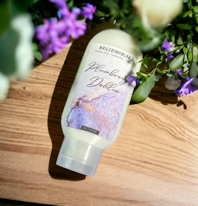 A bottle of Plumberry dahlia hand and body lotion lies on a wooden surface surrounded by purple flowers. The bottle is cream-colored with a purple and pink graphic label.