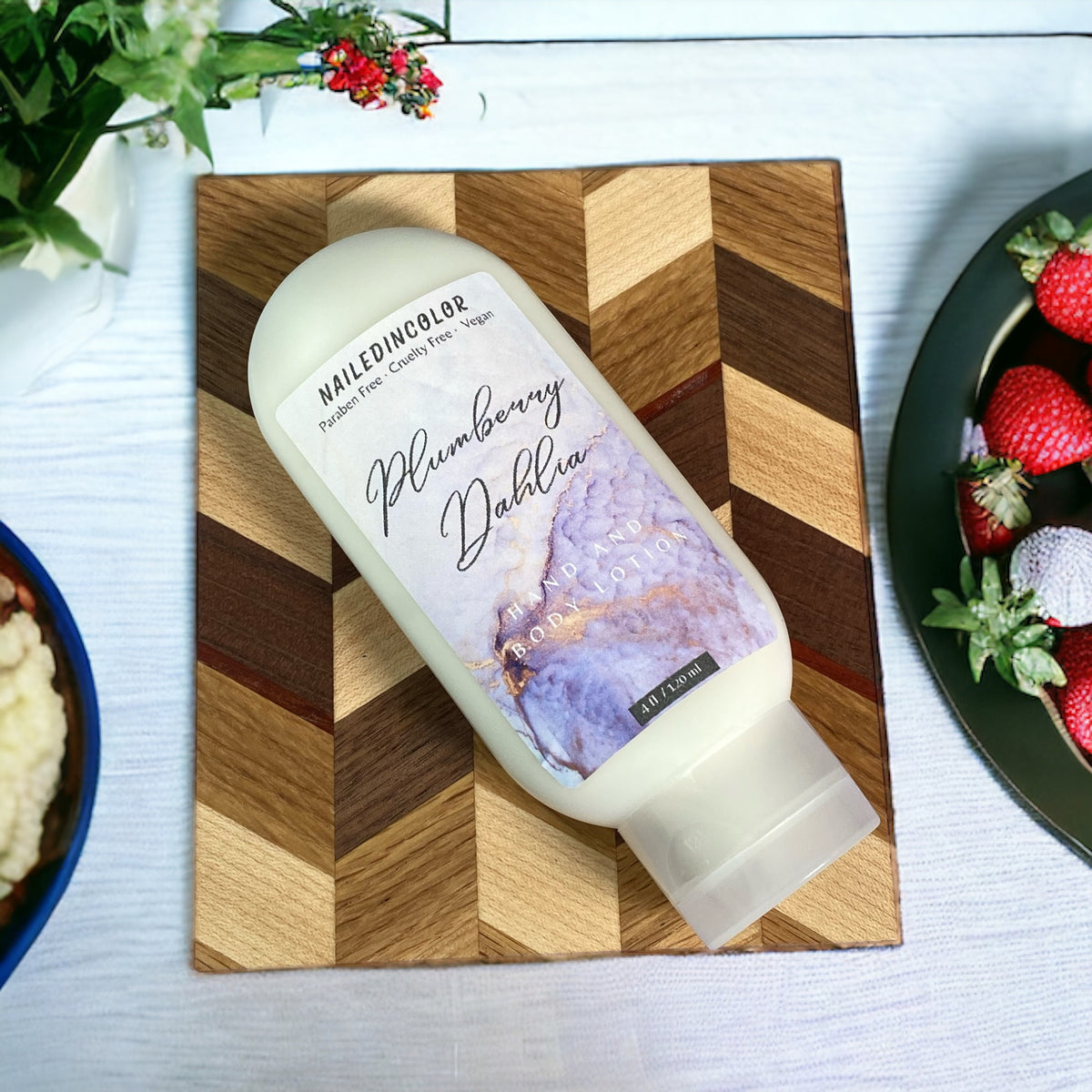 A bottle of NailedinColor Plumberry Dahlia hand cream placed diagonally on a wooden cutting board, surrounded by food props like strawberries and a bowl on a light tabletop.