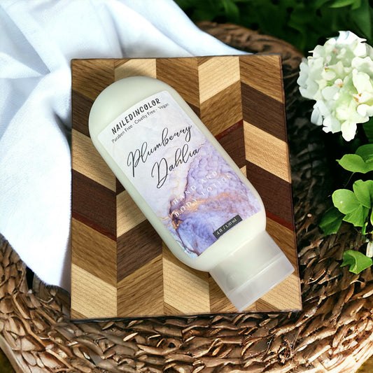 A bottle of Plumberry Dahlia hand and body care lotion placed diagonally on a geometric wooden block, styled on a woven tray with a white cloth and white flowers.