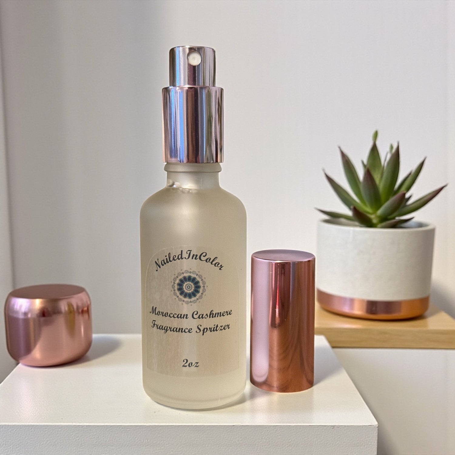Nailed In Color Moroccan Cashmere Fragrance Spritzer bottle with rose gold cap and base on a white surface with a plant in the background.