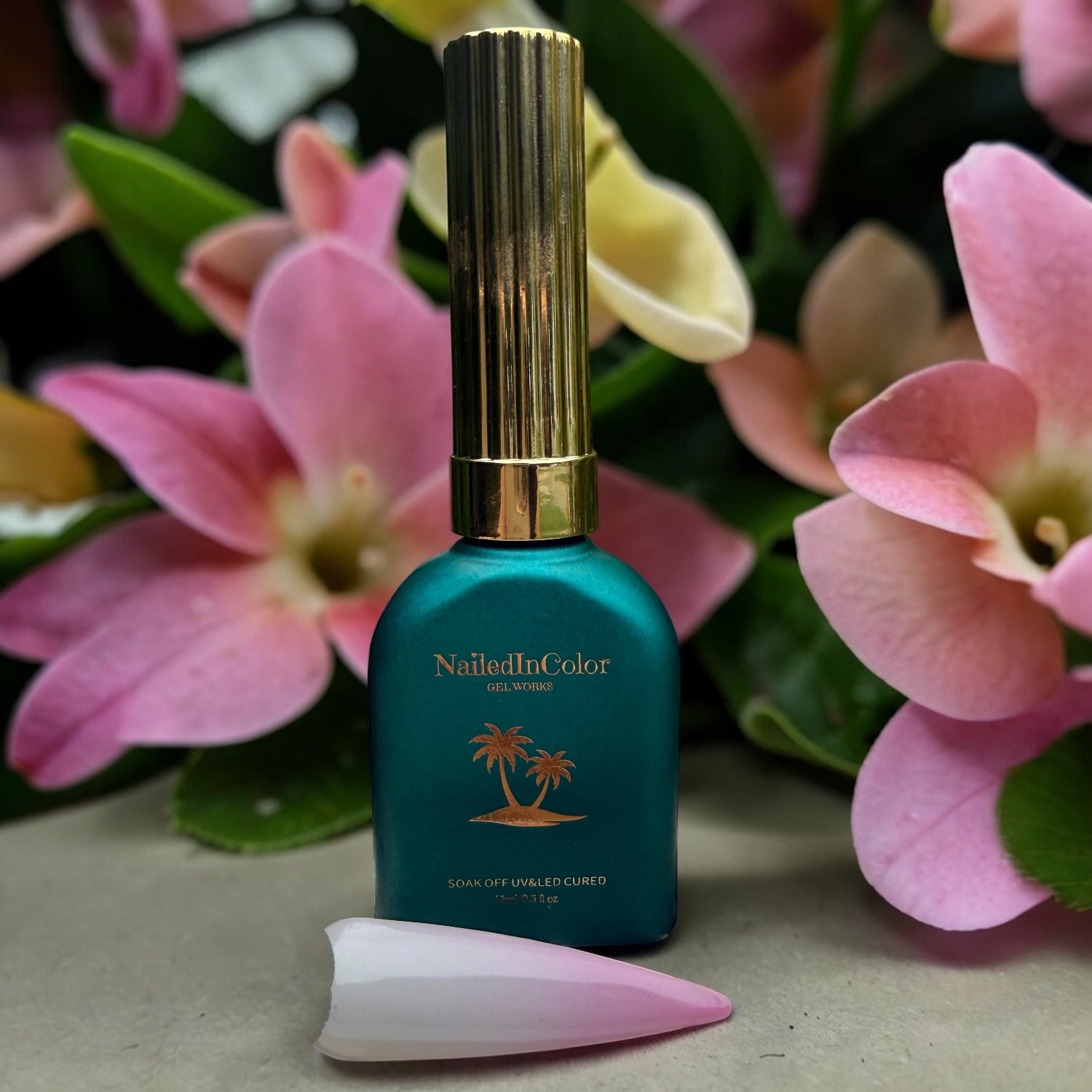 Green gel polish bottle with gold cap in front of flowers and with a swatch of white and light pink