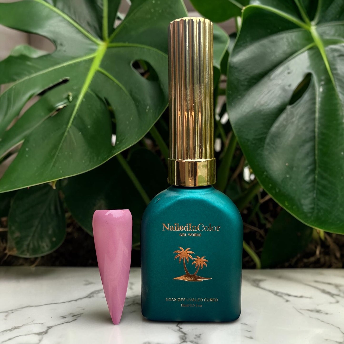 Green gel polish bottle with gold cap in front of green plants and a mauve nail swatch