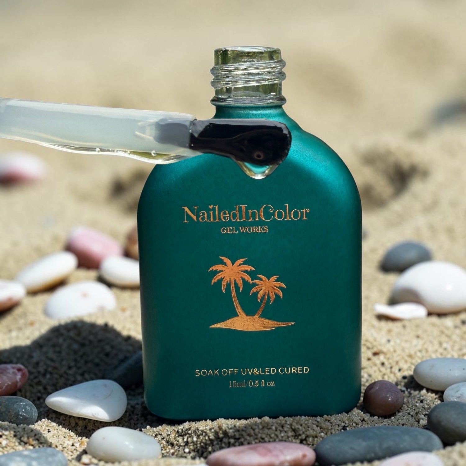 Green NailedInColor gel bottle on sand with pebbles, showing a polish brush holding some gel base.