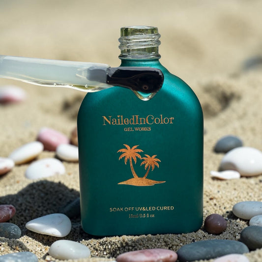 Green NailedInColor gel bottle on sand with pebbles, showing a polish brush holding some gel base.