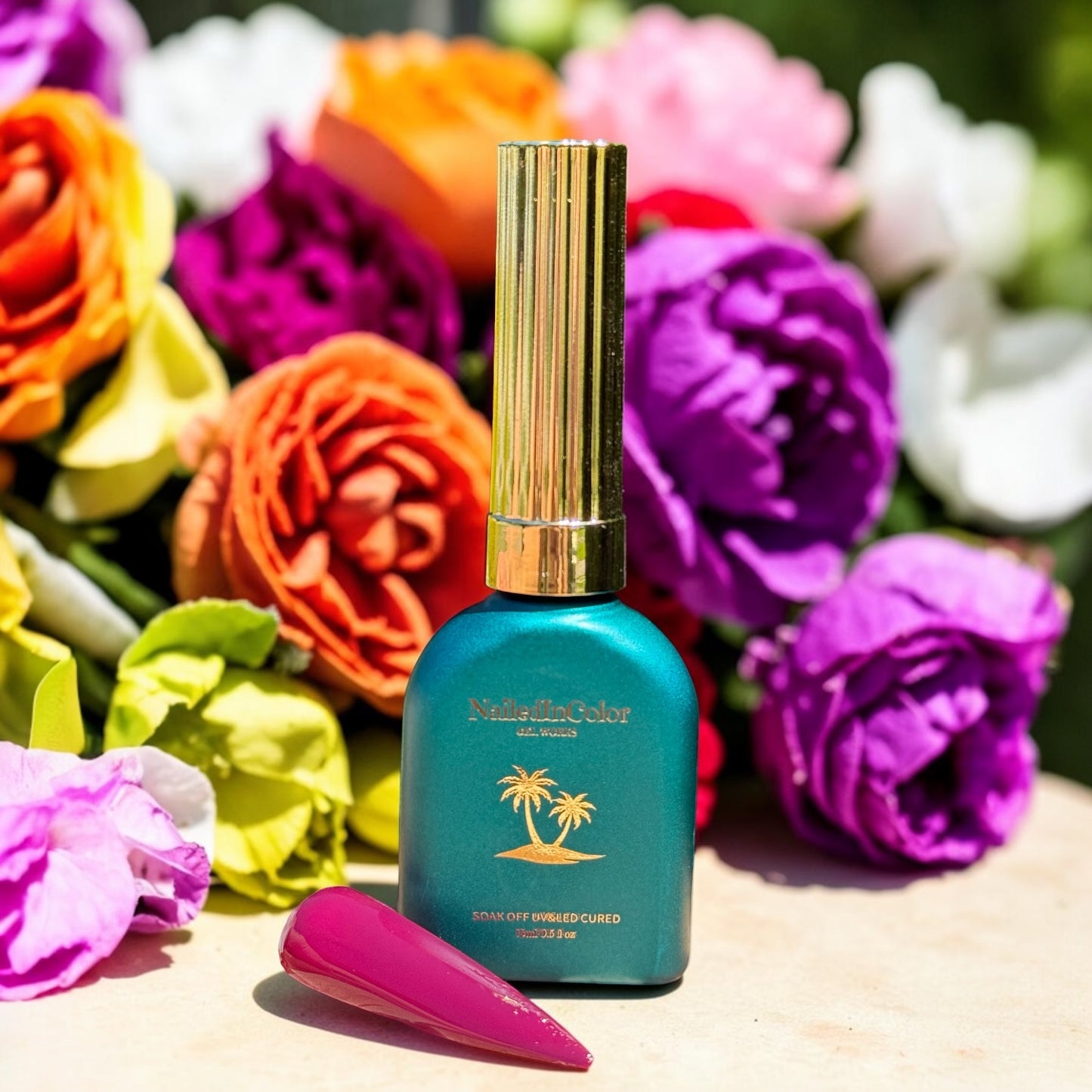 Green nail polish bottle with gold cap on a wooden surface with colorful flowers in the background and a light to dark maroon nail swatch