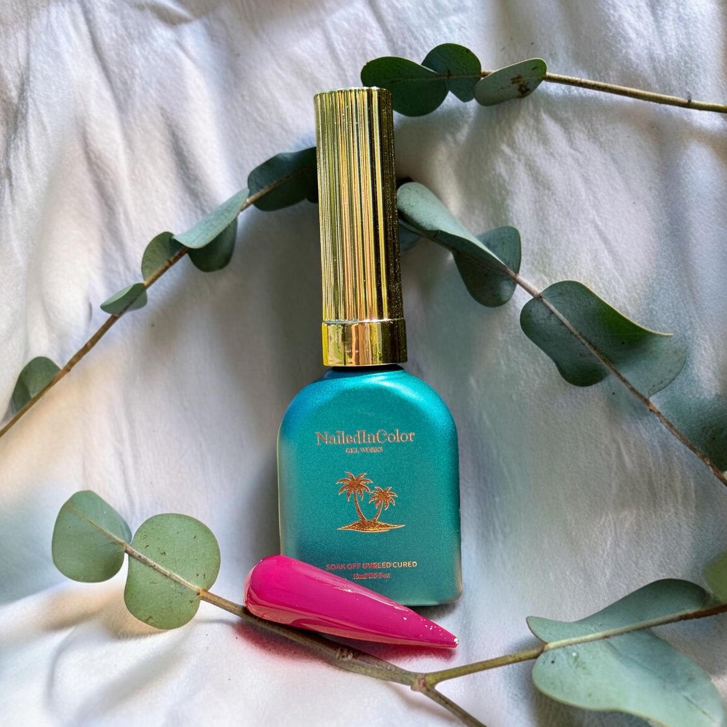 Green nail polish bottle with gold cap on a white fabric background with eucalyptus leaves next to a polished nail swatch in rose to dark maroon