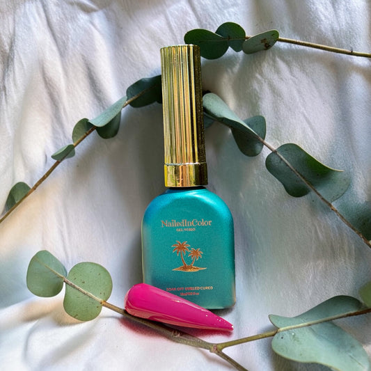 Green nail polish bottle with gold cap on a white fabric background with eucalyptus leaves next to a polished nail swatch in rose to dark maroon