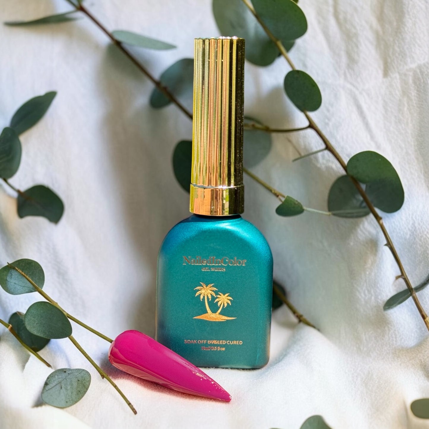 Green  nail polish bottle with gold cap on a white background surrounded by eucalyptus leaves with a light to dark maroon nail polish swatch