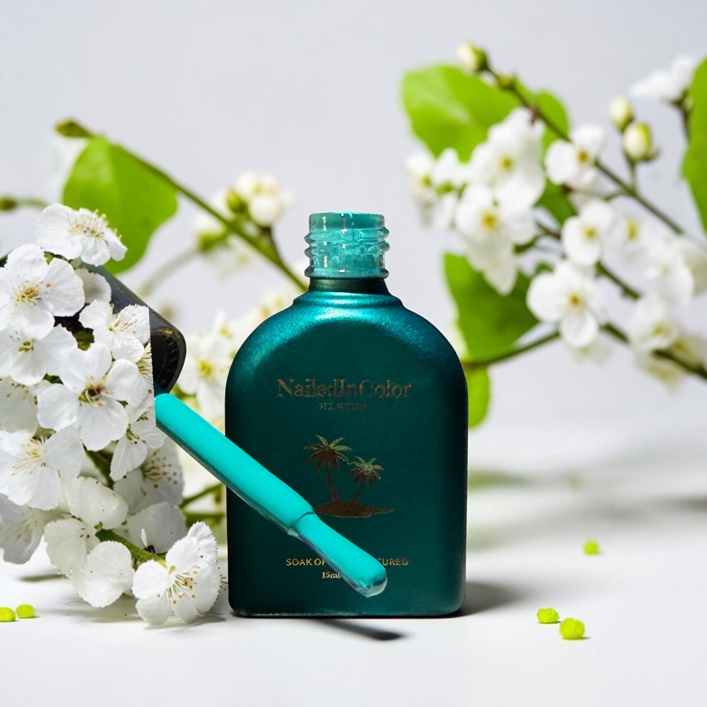 A green gel polish bottle with a polish brush dipped in the blue green shade surround by small white flowers on a counter