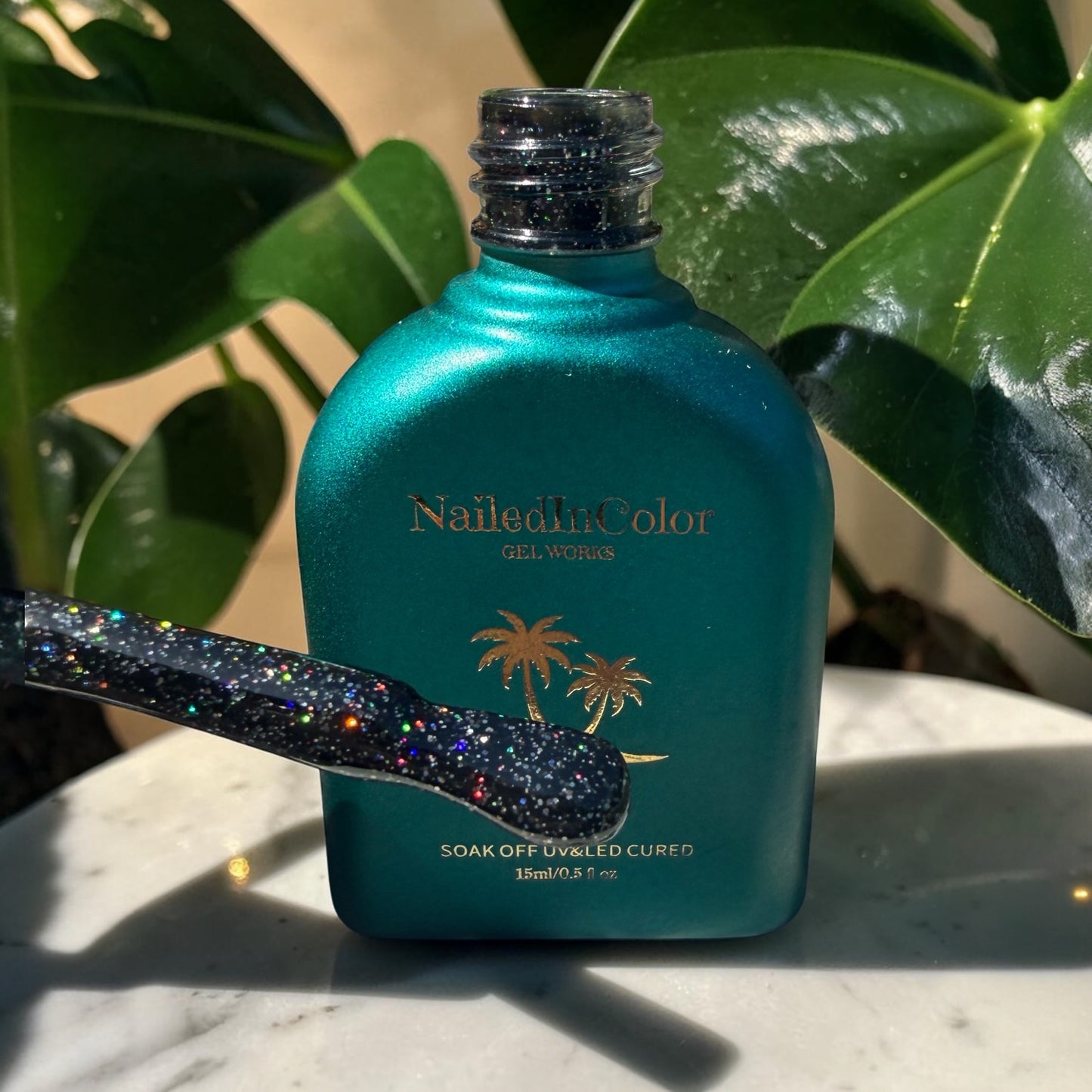 Green bottle of NailedinColor gel polish with a glittery brush on a marble surface with green leaves in the background