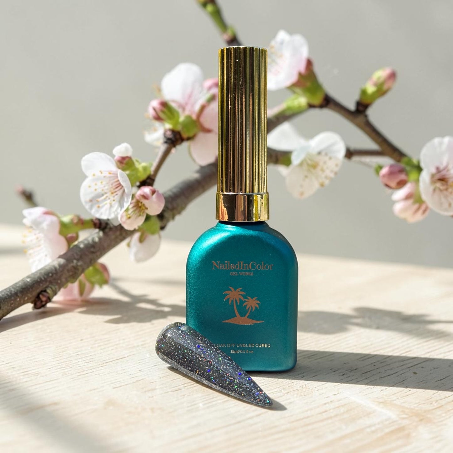 Teal nail polish bottle with gold cap and a nail swatch with black holographic polish on a wooden surface with cherry blossom branch in the background