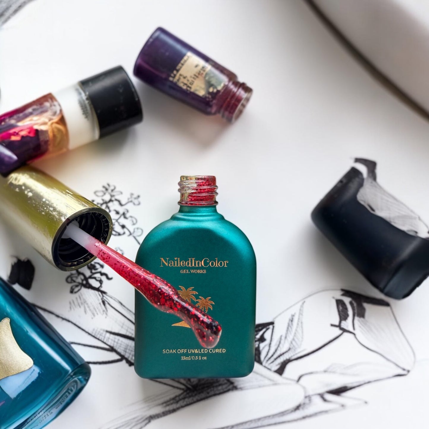 Nail polish bottles with a focus on a polish brush with red glitter polis on it next to a teal bottle labeled 'NailedInColor' on a white surface with drawings