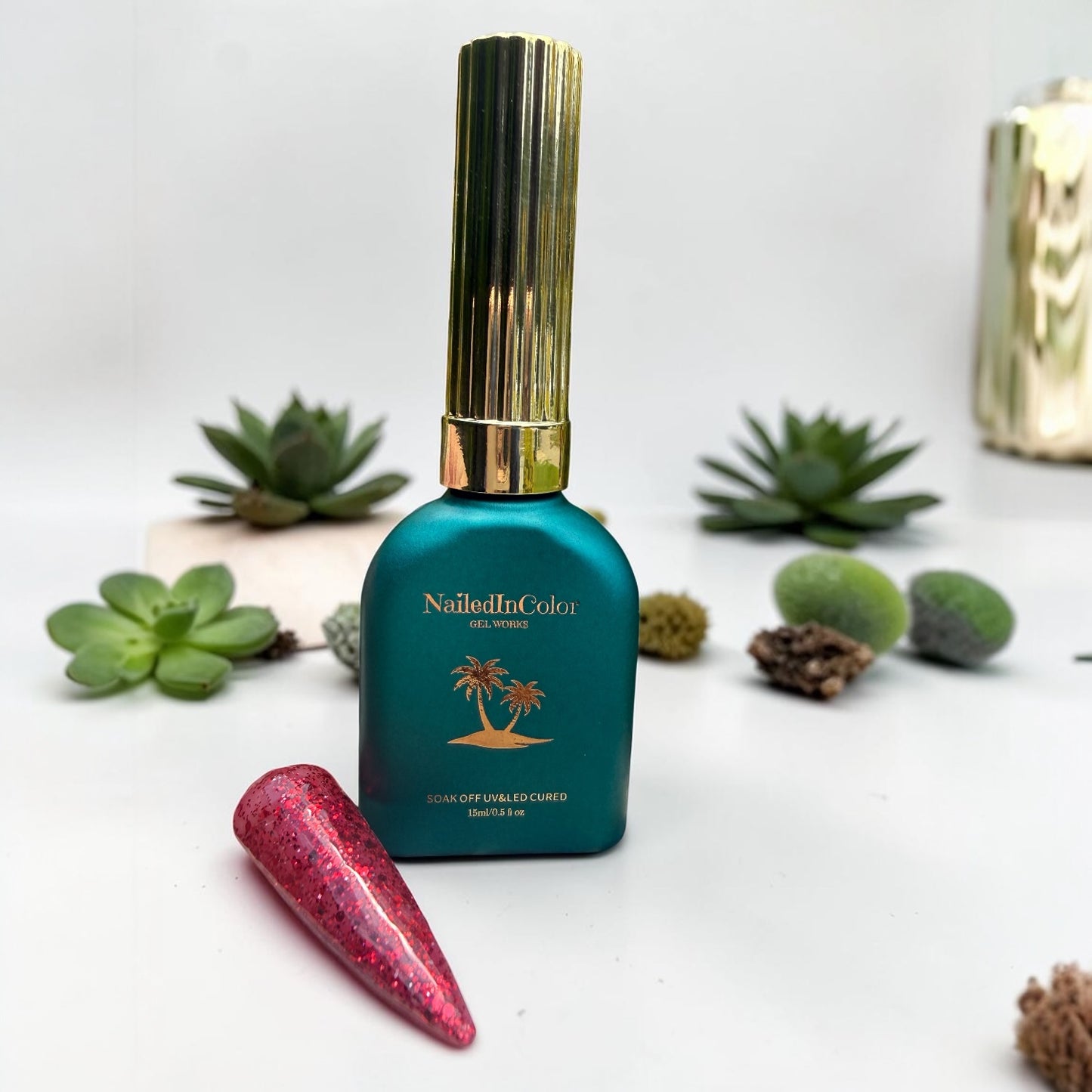 Nail polish bottle with gold cap and red glittery nail swatch on a white background with succulents.