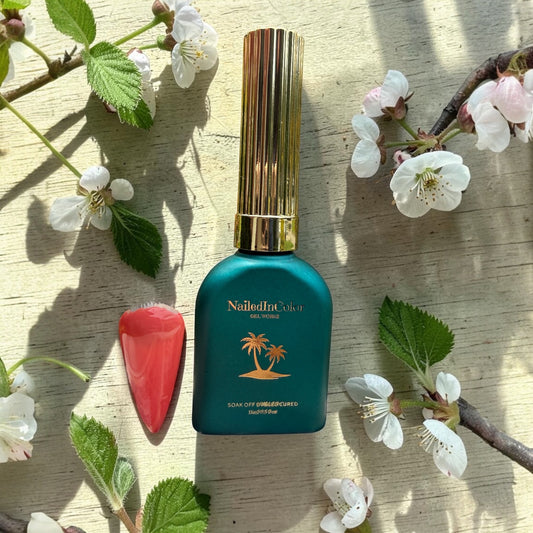 Green nail polish bottle with gold cap on a wooden surface with cherry blossoms and a coral polished nail swatch
