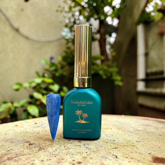 Green gel polish bottle with gold cap on a table with florals and greenery in background. Cat eye medium blue sparkle nail sample next to bottle