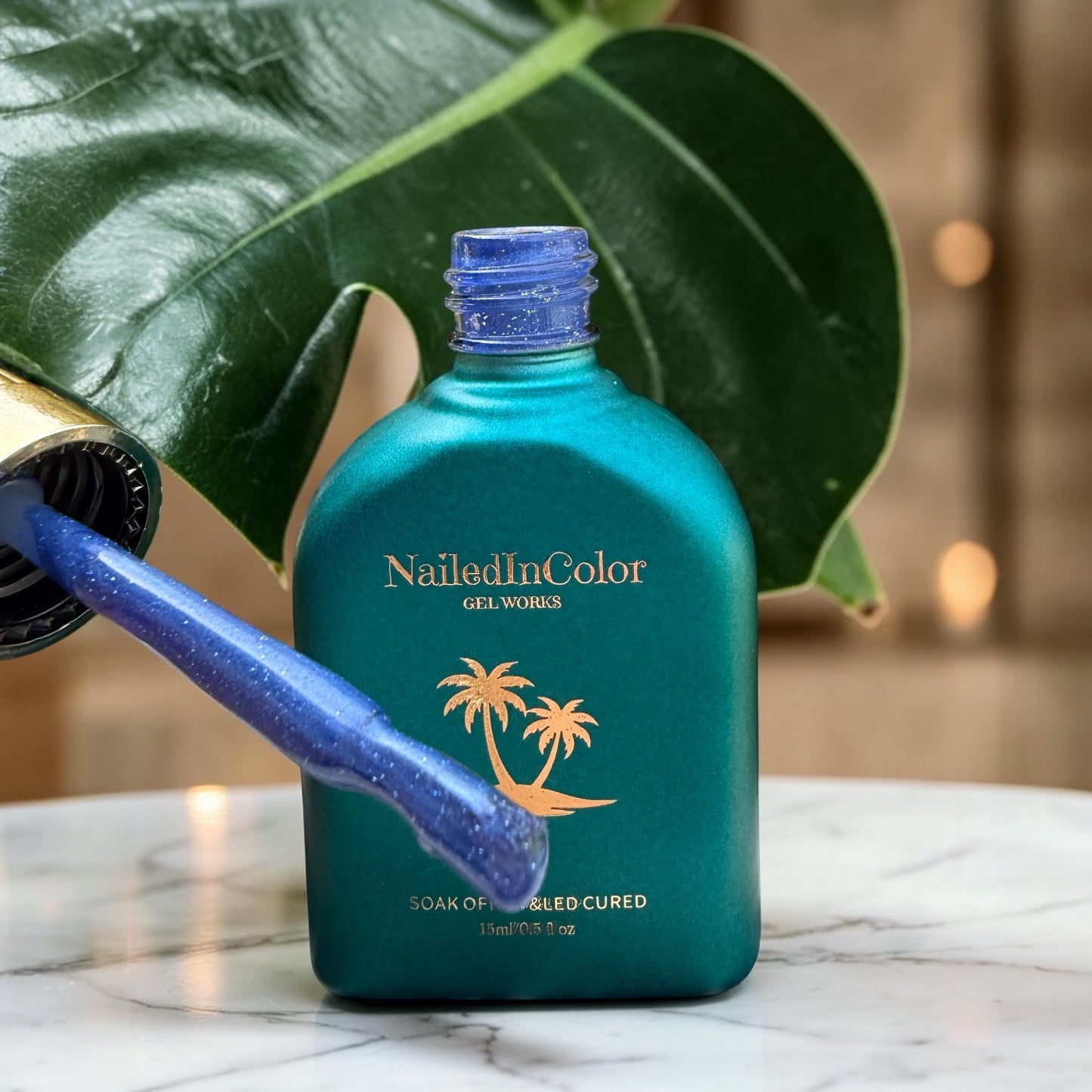Green bottle of NailedInColor gel with palm tree design on a marble surface with green leaves in the background and a brush applicator with sparkly blue gel polish on it