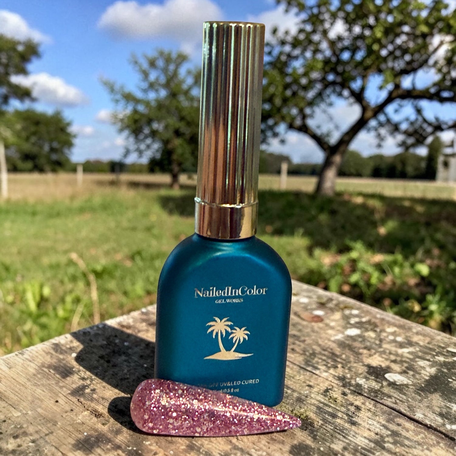 Green nail polish bottle with gold cap on a wooden surface with a pink and silver glitter nail swatch outdoors