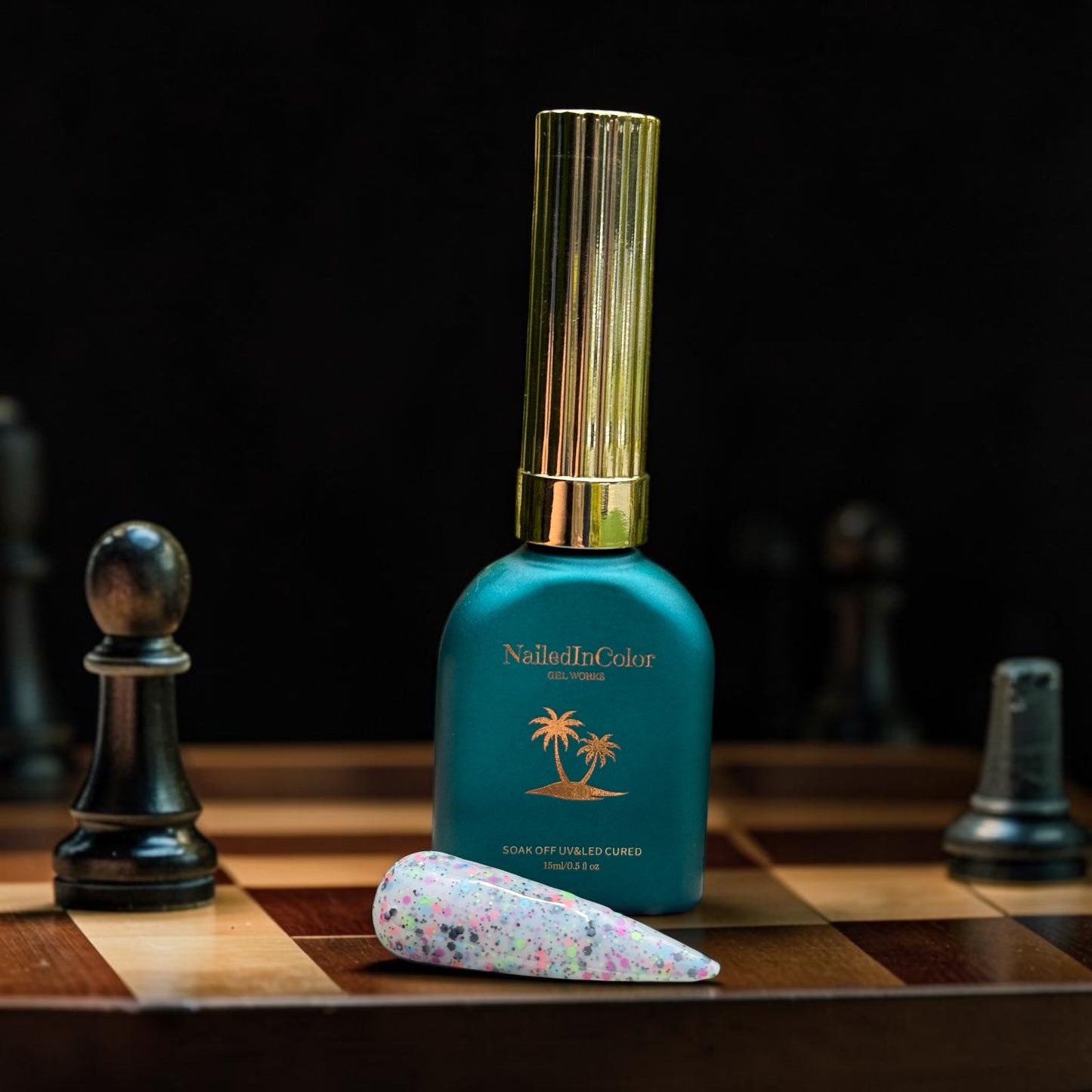 Green nail polish bottle with gold cap on a chessboard with chess pieces and a nail swatch done in Black Tie Confetti gel polish.black flake with various other small flakes on a cream background.