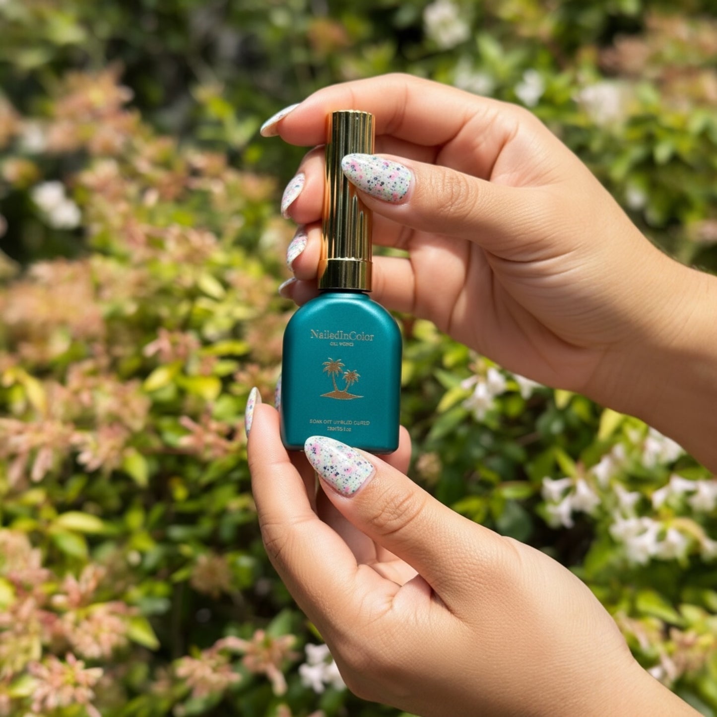Hand holding a green  bottle with a gold cap against a green foliage background nails painted in black speckled gel polish called Black Tie Confetti