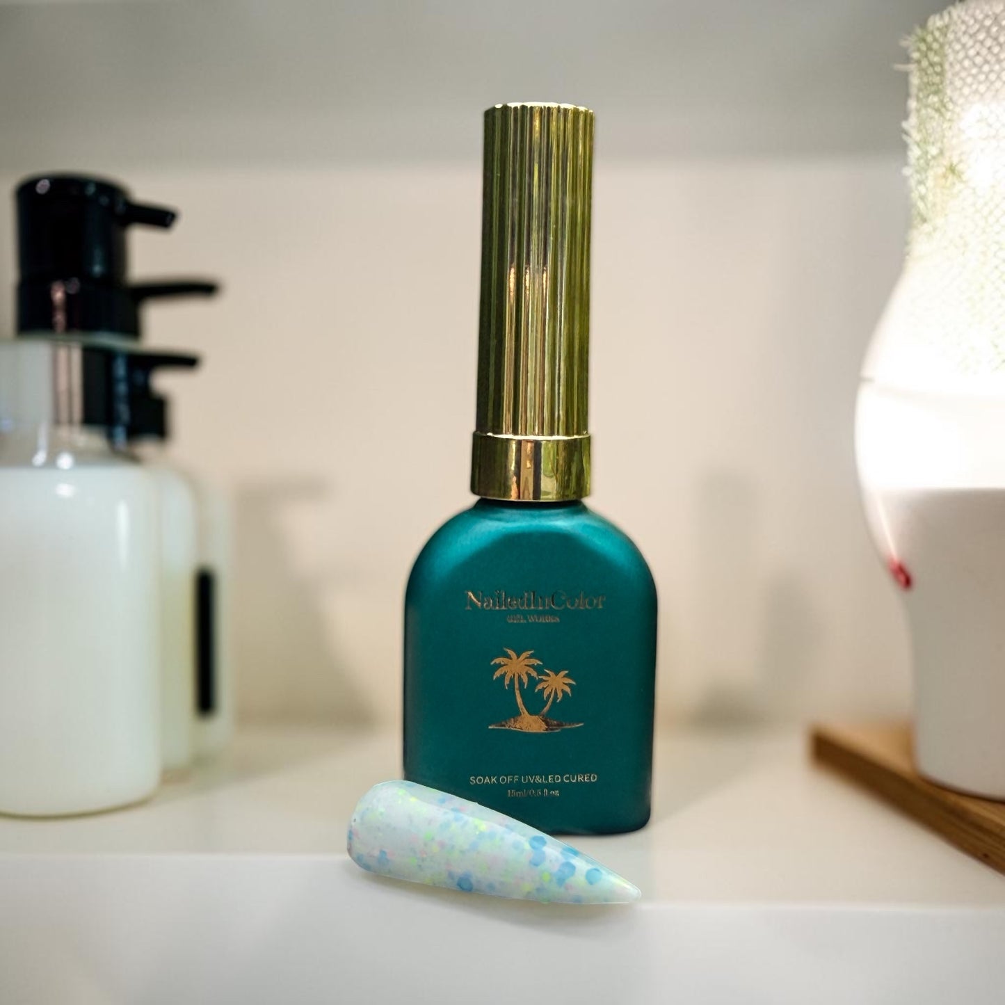 Teal nail polish bottle with gold cap on a bathroom counter with pump bottles and a polished nail swatch with the multicolor blue flakes