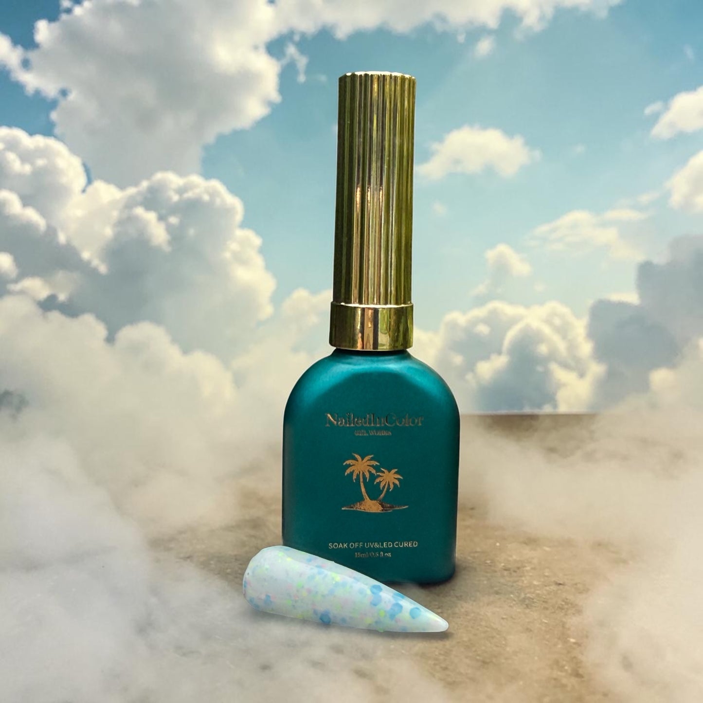 Teal nail polish bottle with gold cap on sand with clouds in the background and wisps of fog, multicolor flake polished nail swatch