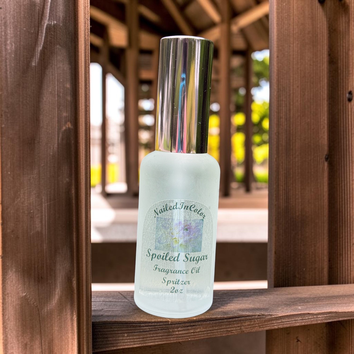 Frosted bottle of 'Spoiled Sugar' fragrance oil spritzer on a wooden surface with blurred outdoor background