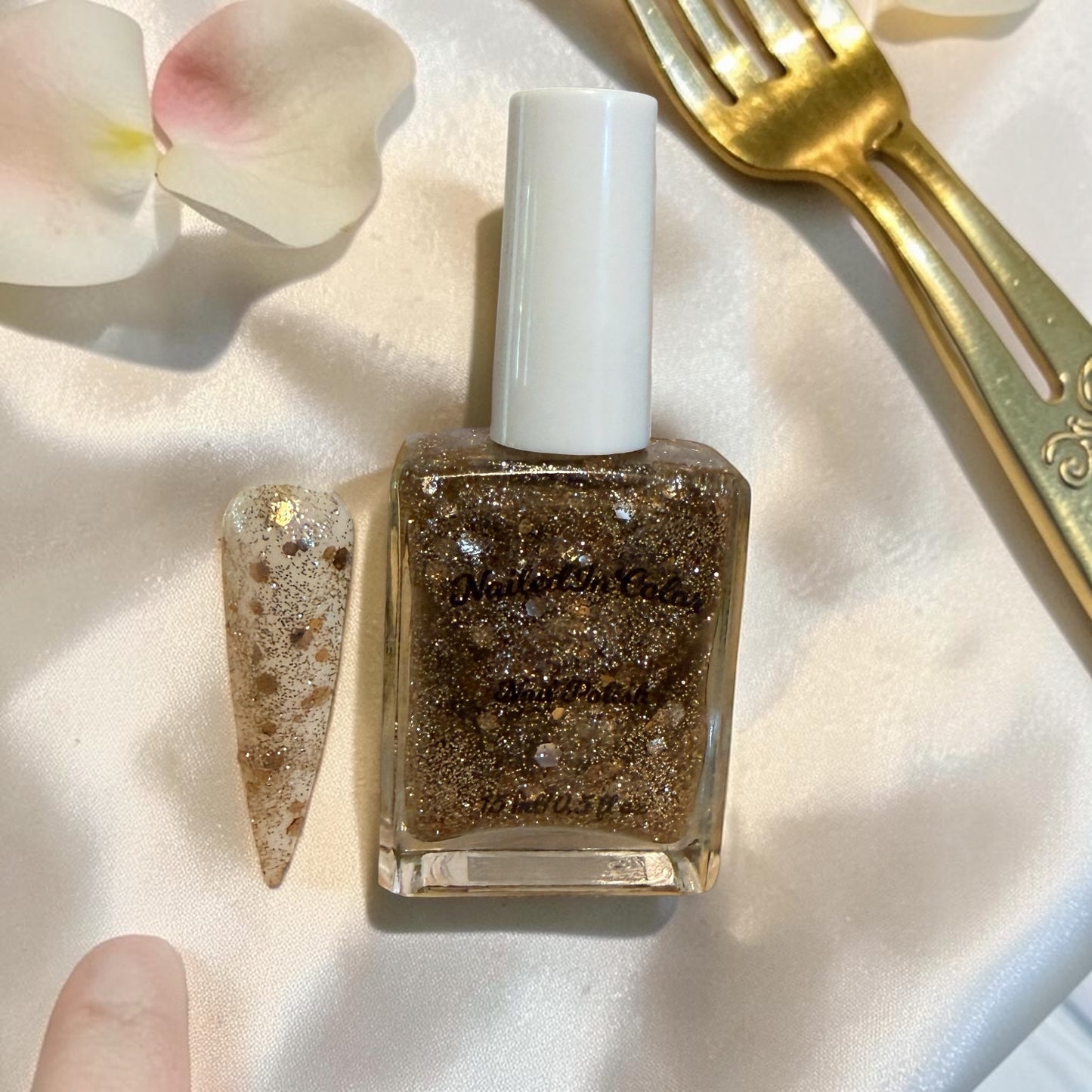 Gold glitter nail polish bottle with a white cap on a light fabric background with a gold glitter nail swatch.
