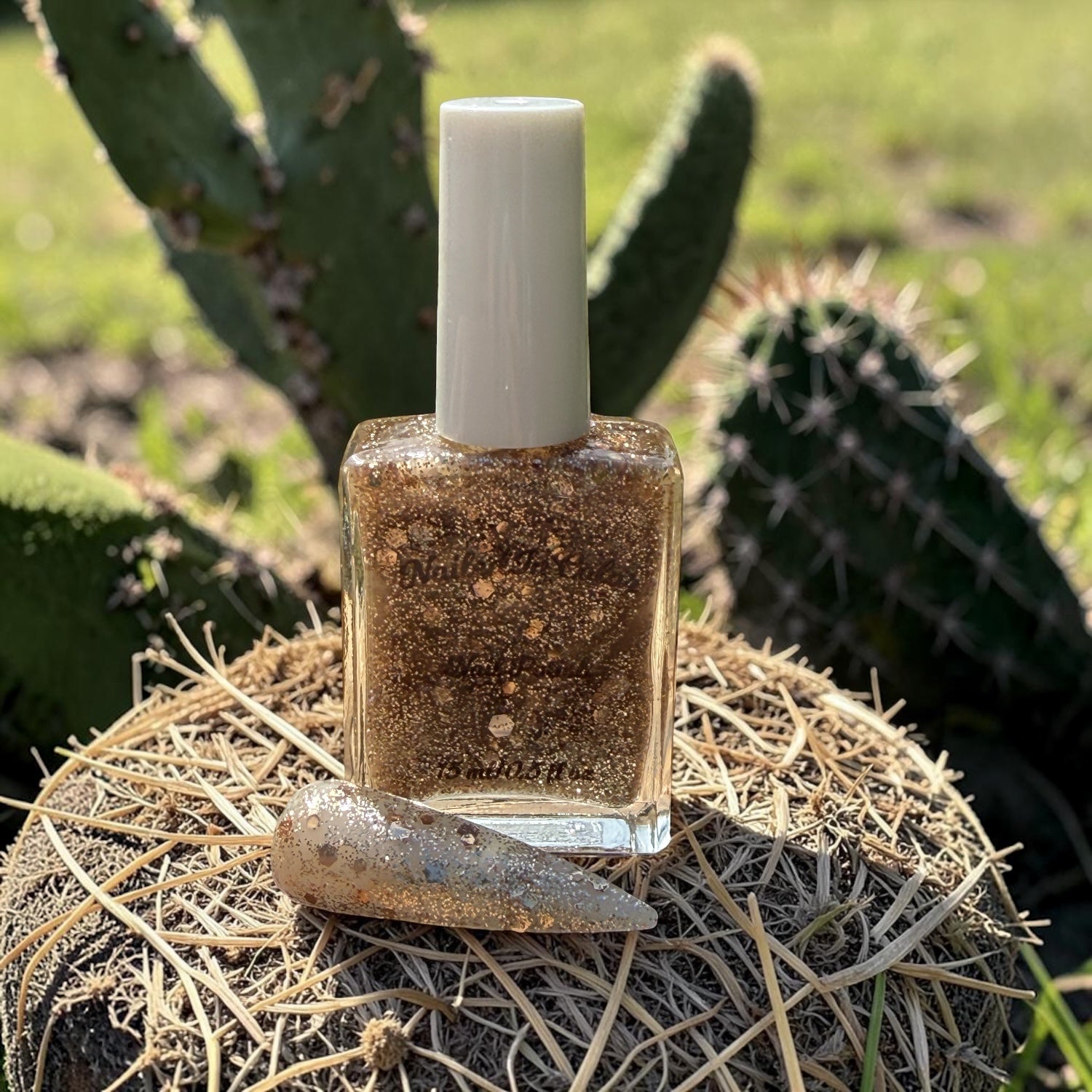 Nail polish bottle with white cap and a matching nail swatch on straw in front of a cactus plant.