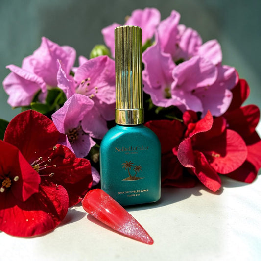 Green nail polish bottle with gold cap on a white surface surrounded by pink and red flowers, red nail tip with fine silver glitter cat eye topcoat