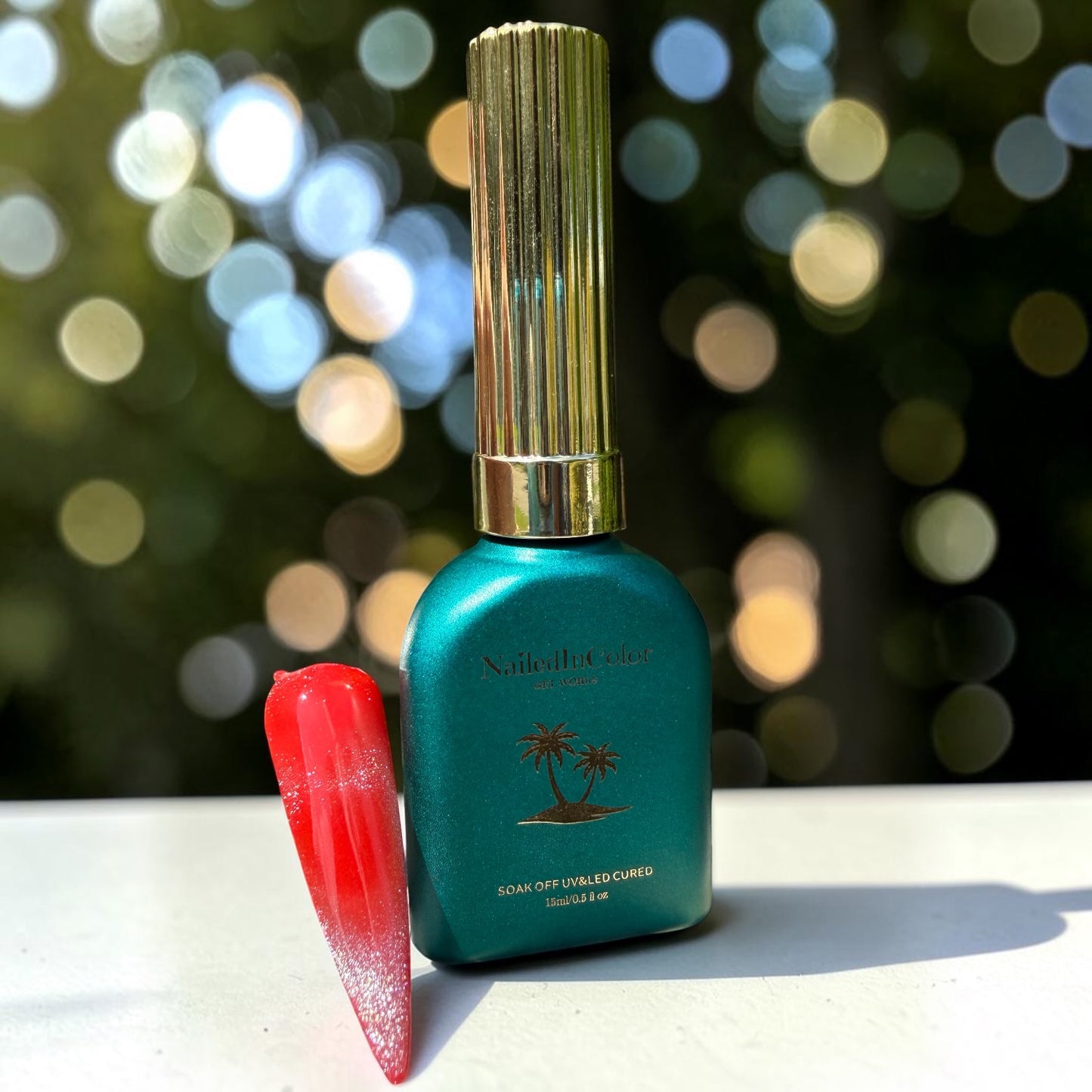 Teal nail polish bottle with gold cap and red nail tip, silver glitter cat eye topcoat on a blurred bokeh background