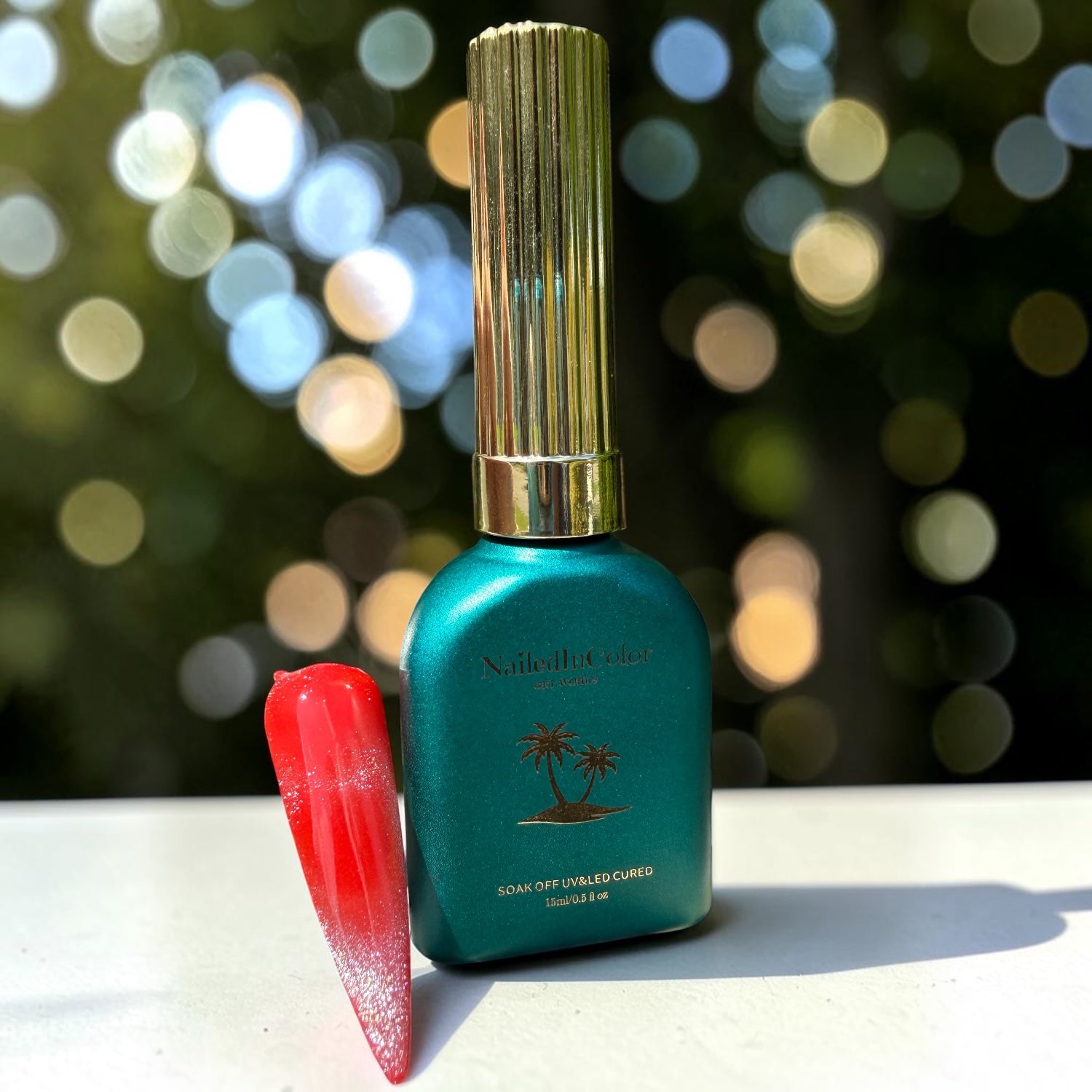 Teal nail polish bottle with gold cap and red nail tip, silver glitter cat eye topcoat on a blurred bokeh background