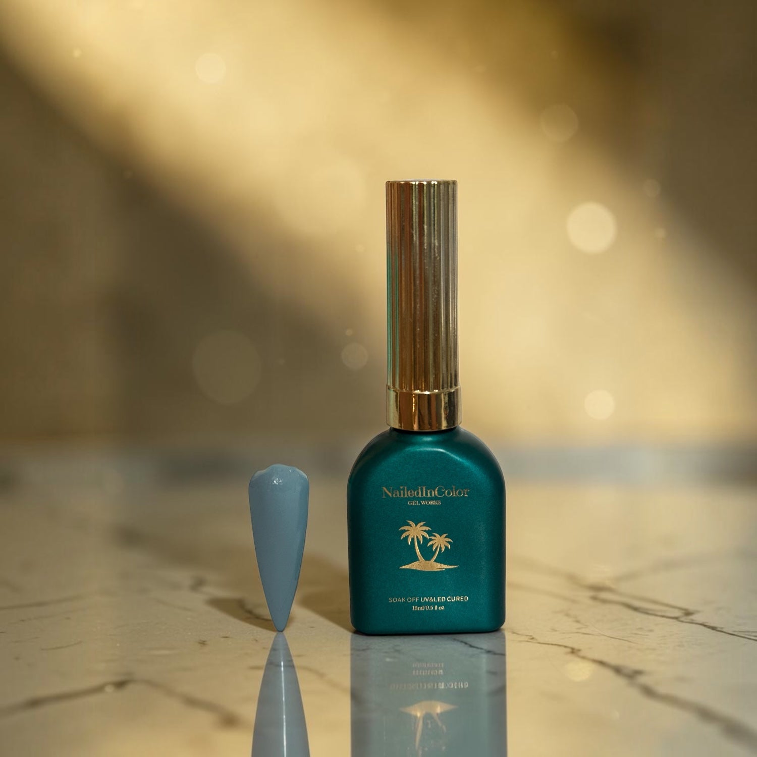 teal green nail polish bottle with gold cap on a marble counter and gold sparkle in background and a blue nail tip