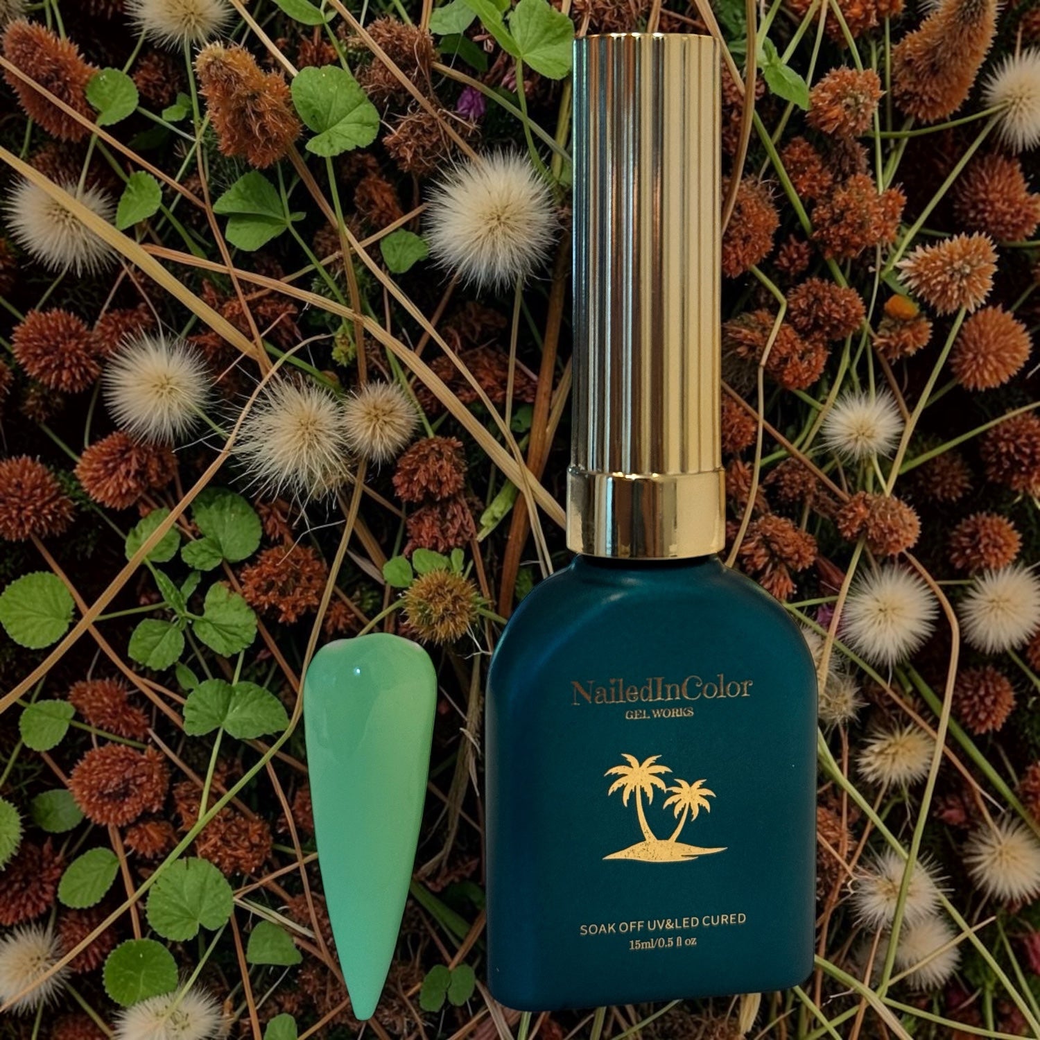 Teal green polish bottle with gold cap laying on florals and greens with a light green nail tip