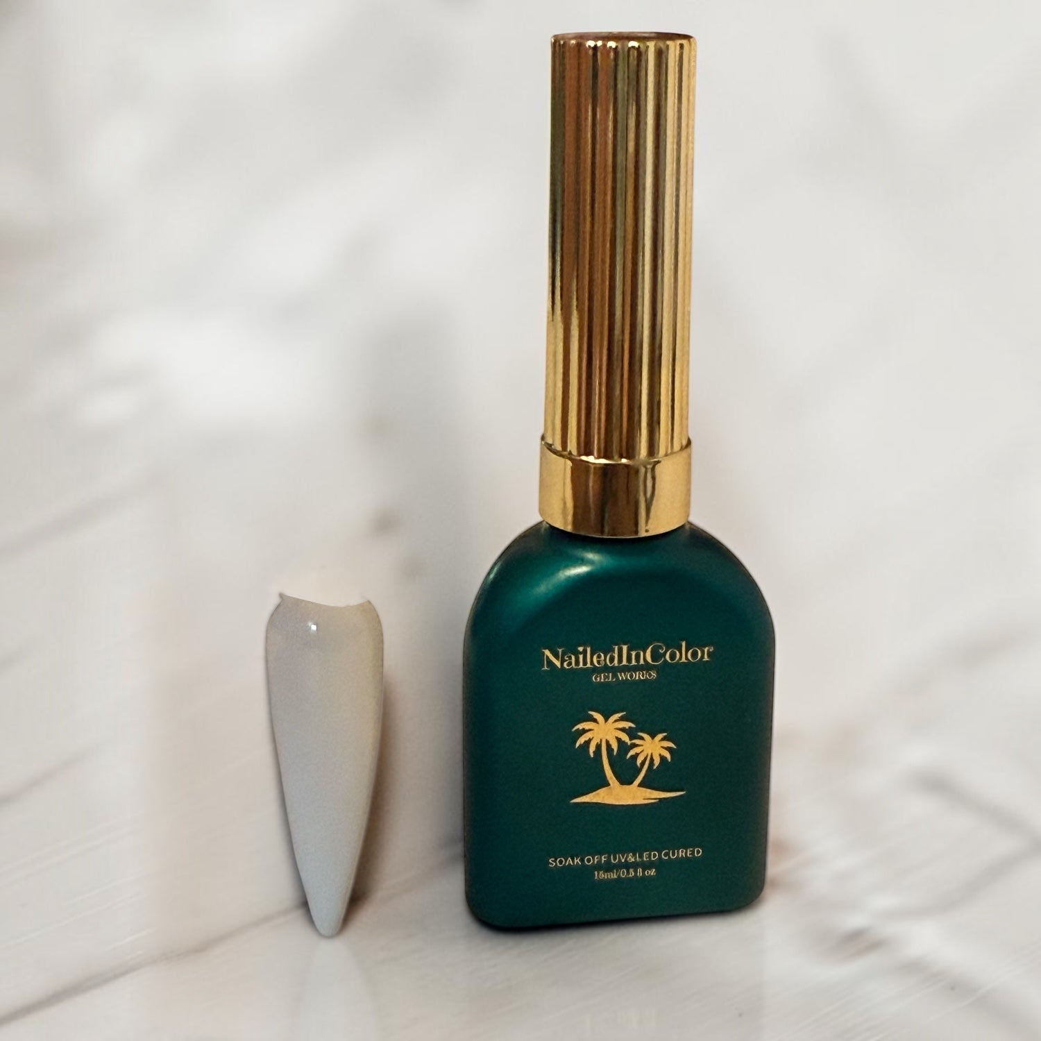 white gel polish nail tip against a gray marble wall and base alongside a green polish bottle with a gold cap