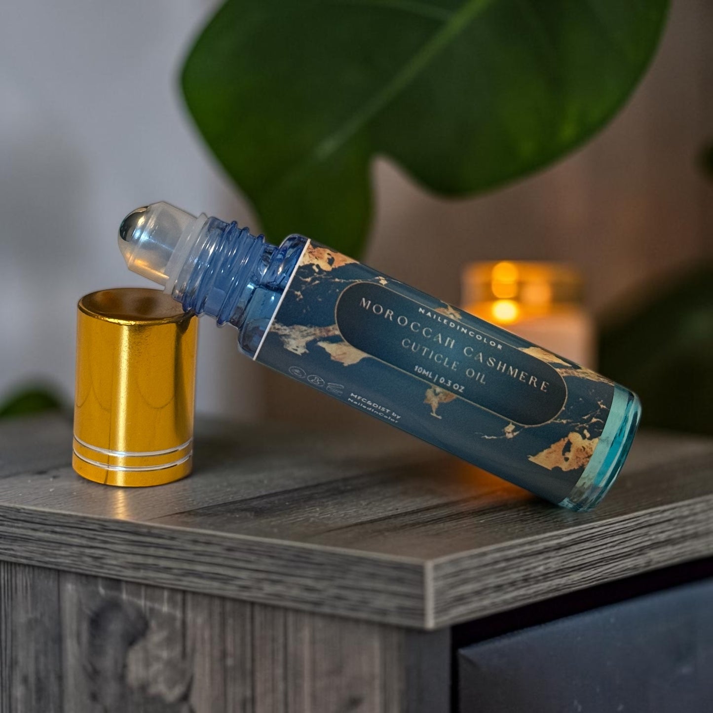 A bottle of Moroccan  Cashmere Cuticle Oil with a blue label and gold map pattern, shown with its gold cap off and resting beside it on a wooden surface with a candle and blurred background.