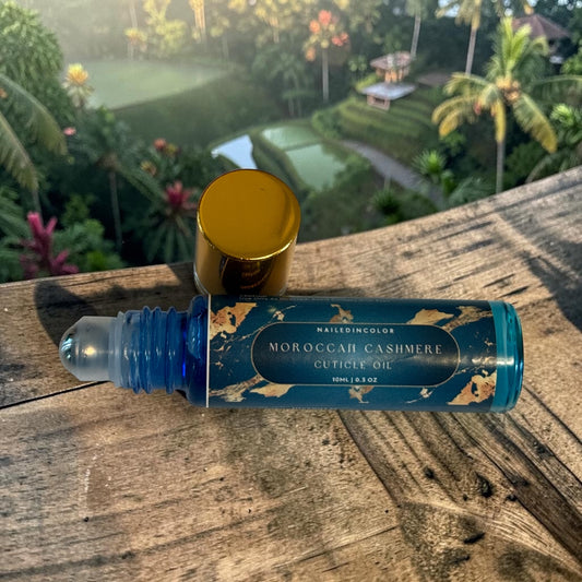A roll-on bottle of Moroccan Cashmere Cuticle Oil with a blue label and gold cap, resting on a rustic wooden surface with a tropical garden background.