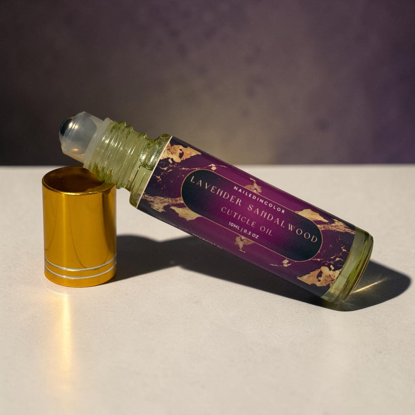 A bottle of Lavender Sandalwood Cuticle Oil with its gold cap removed and placed beside it on a light surface, set against a dark purple background.