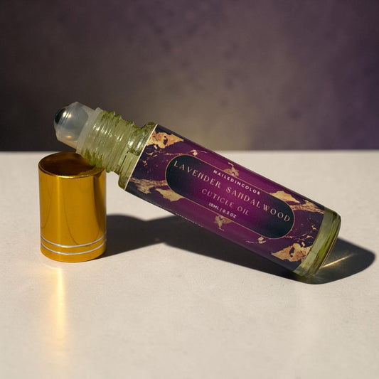A bottle of Lavender Sandalwood Cuticle Oil with its gold cap removed and placed beside it on a light surface, set against a dark purple background.