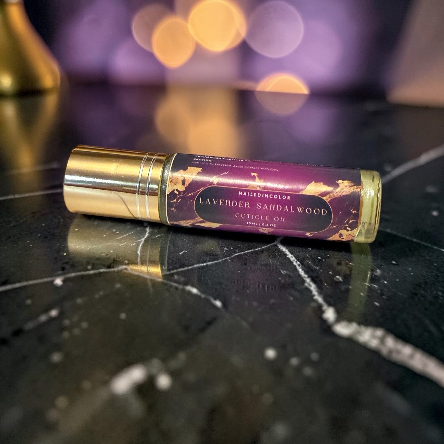 A nail color rollerball bottle labeled  Lavender Sandalwood Cuticle Oil' resting on a polished black marble surface with a blurred purple and gold bokeh background.