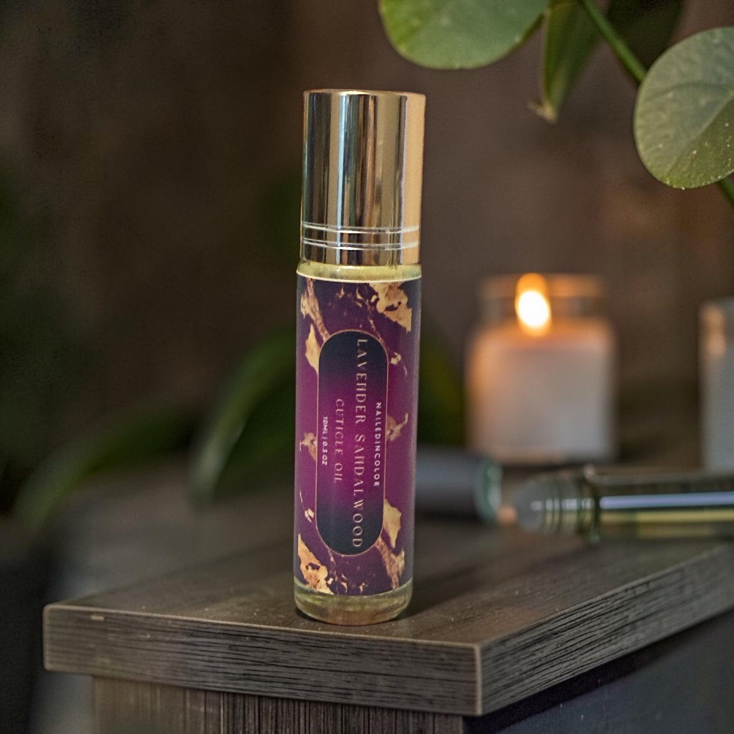 A clear glass roll-on bottle with a metallic gold cap and a purple label reading Lavender Sandalwood standing on a dark wooden surface with a softly blurred candle and green leaves in the background.